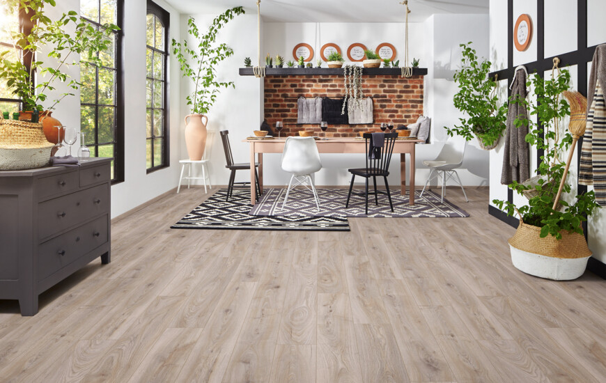 Dining room with light laminate flooring, featuring a rustic brick wall, plants, and a mix of chairs around a wooden table.